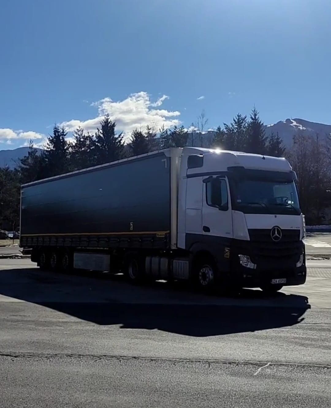 White Mercedes-Benz Actros with a dark curtain trailer and mountains behind